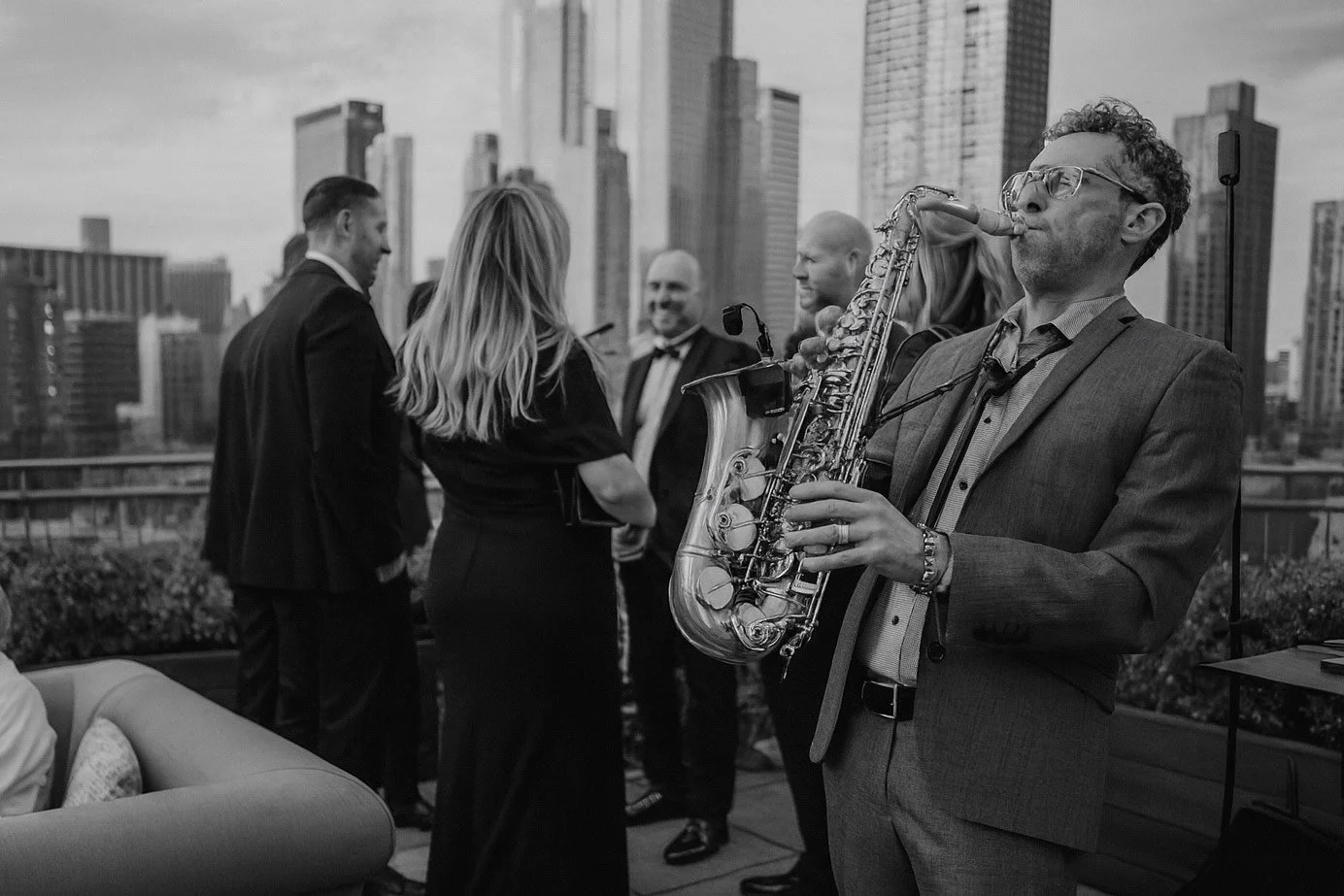 A man in a suit and sunglasses playing a saxophone at an indoor gathering with people socializing in the background.