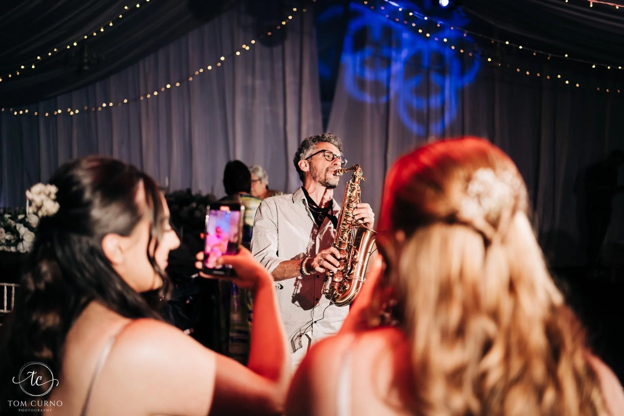 A man playing a saxophone at a festive event with guests watching, some using their phones to take photos, under string lights and decorated drapes.