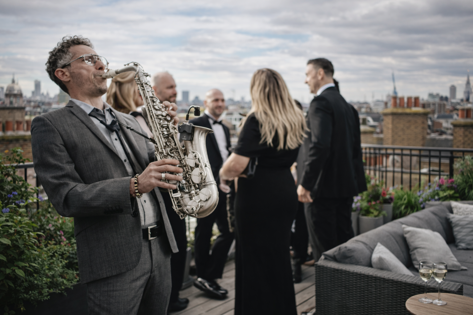 A man in a suit and sunglasses playing a saxophone at an indoor gathering with people socializing in the background.