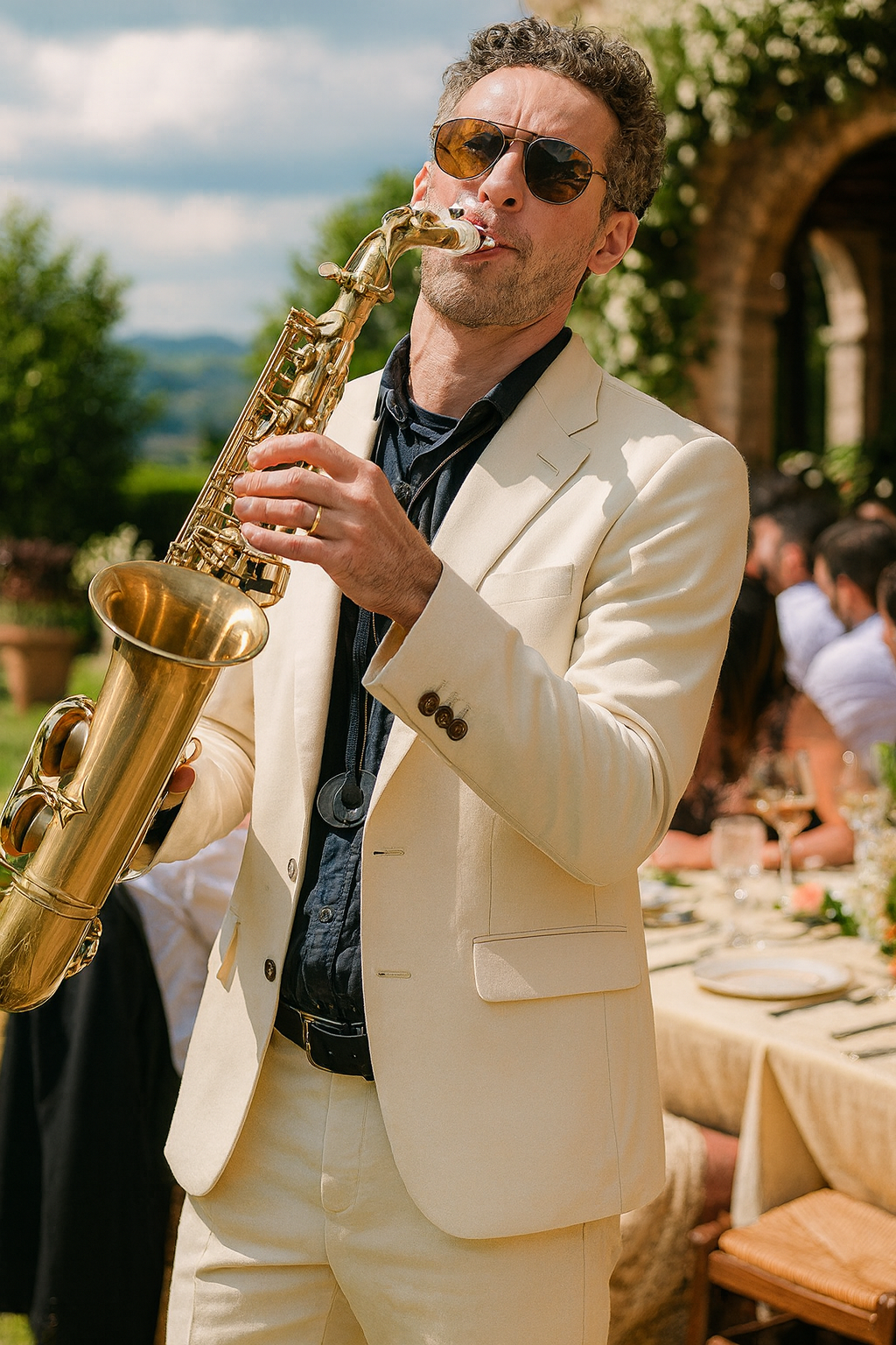 A man wearing sunglasses and a white suit jacket playing a saxophone at an outdoor event.