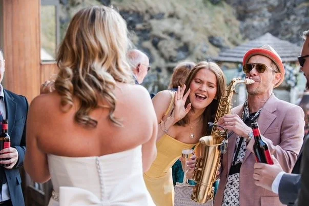 Group of people at an outdoor party, with a woman talking into a microphone, a man playing saxophone, and others holding drinks