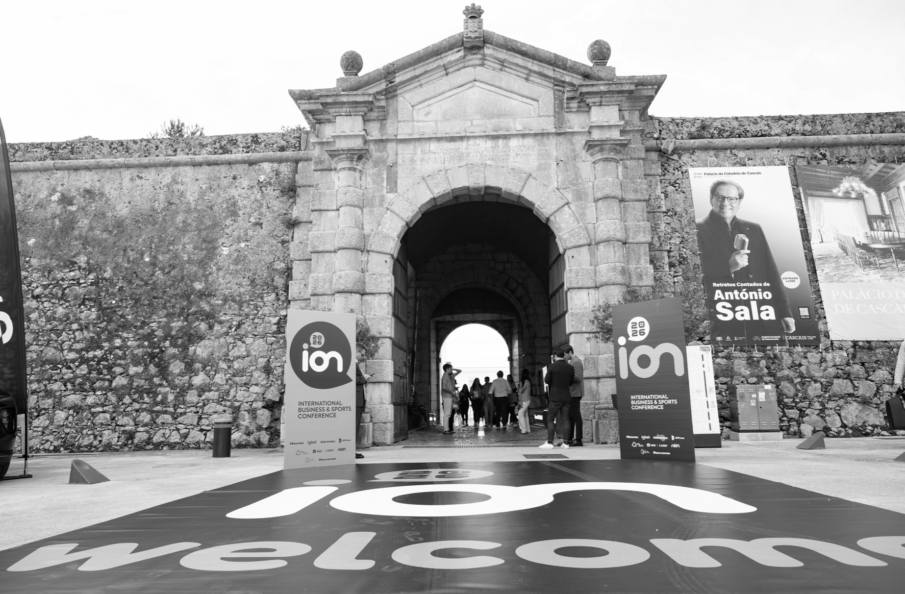 Entrance to a historic stone gate with banners advertising the 2026 International Business & Sports Conference, featuring a photo of António Sala, inside an old city wall with people walking through.