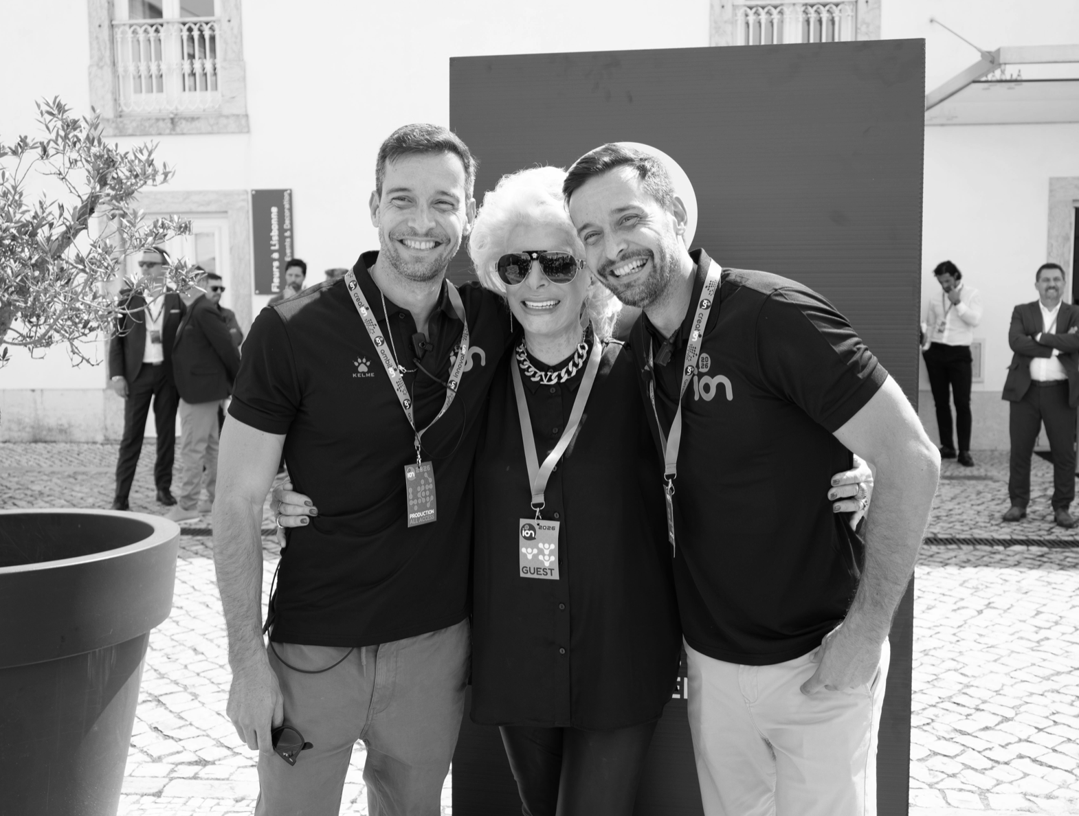 Three smiling people, two men and a woman, standing close together at an outdoor event, with the woman wearing sunglasses and a necklace, and all three wearing event lanyards.