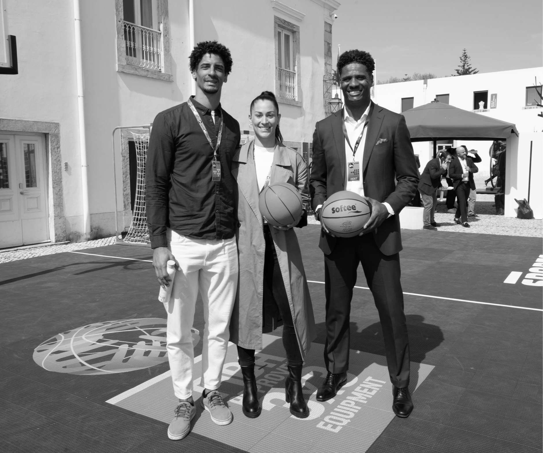 Three people standing together outdoors, holding basketballs, with a building and a group of people in the background.