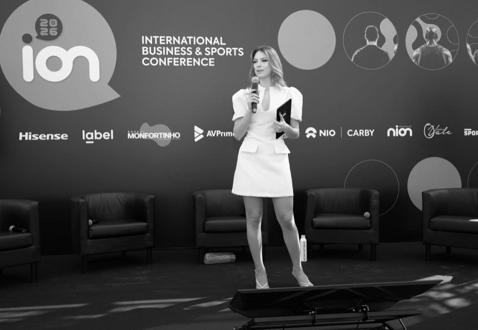 A woman standing on stage holding a microphone and a tablet at the International Business & Sports Conference, with a dark backdrop featuring logos of sponsors and the conference name.