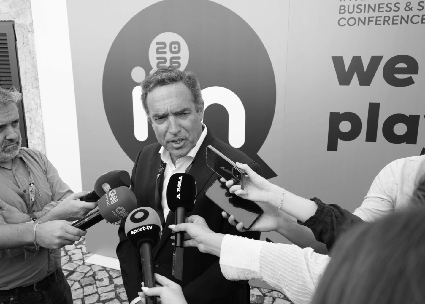 A man in a suit speaking to the press surrounded by microphones from various media outlets, with a background displaying the LinkedIn logo and part of a phrase that reads "we p..." in a conference or event setting.