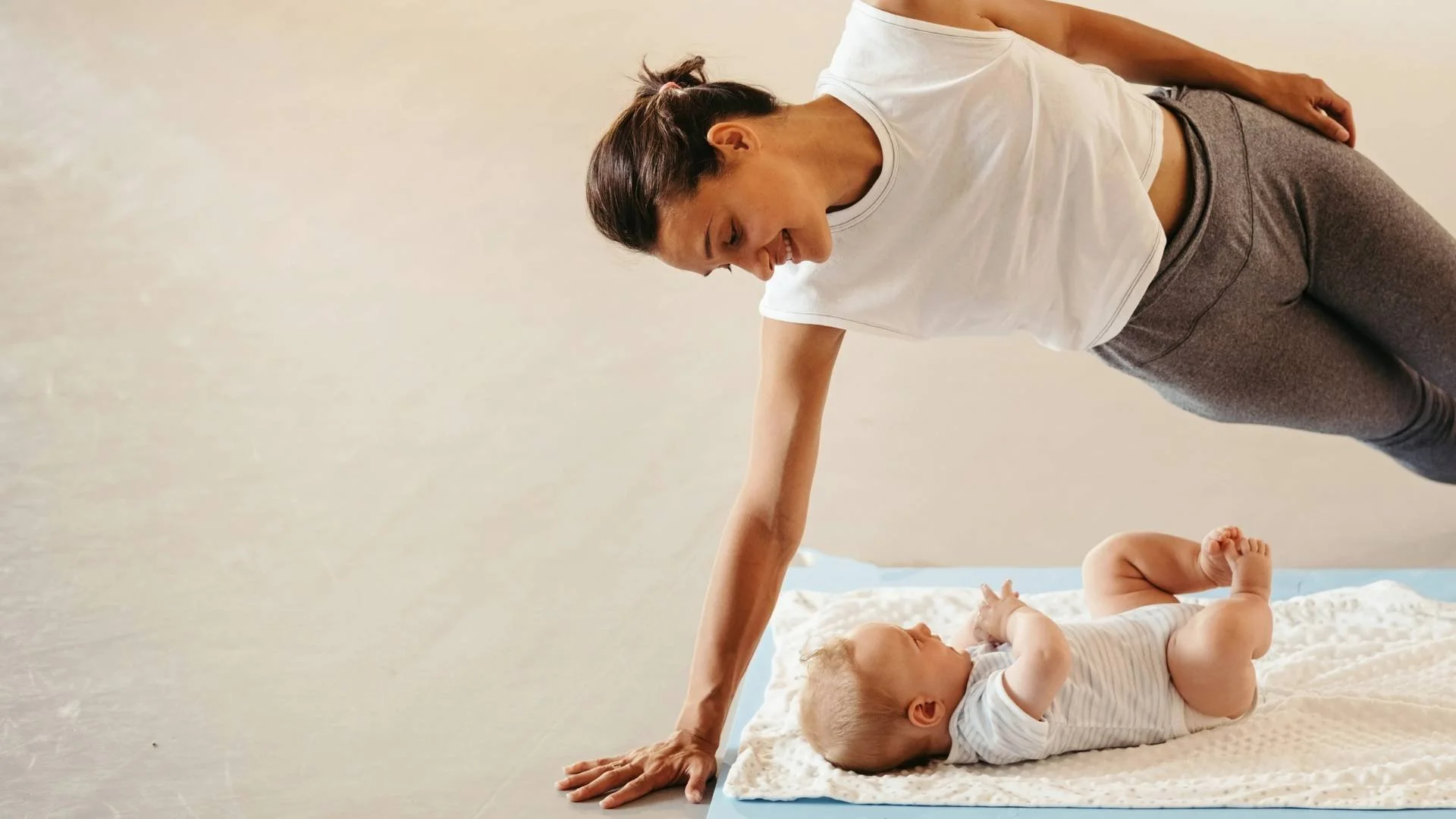 A woman doing a Postnatal Pilates side plank exercise, smiling down at her baby lying beside her.