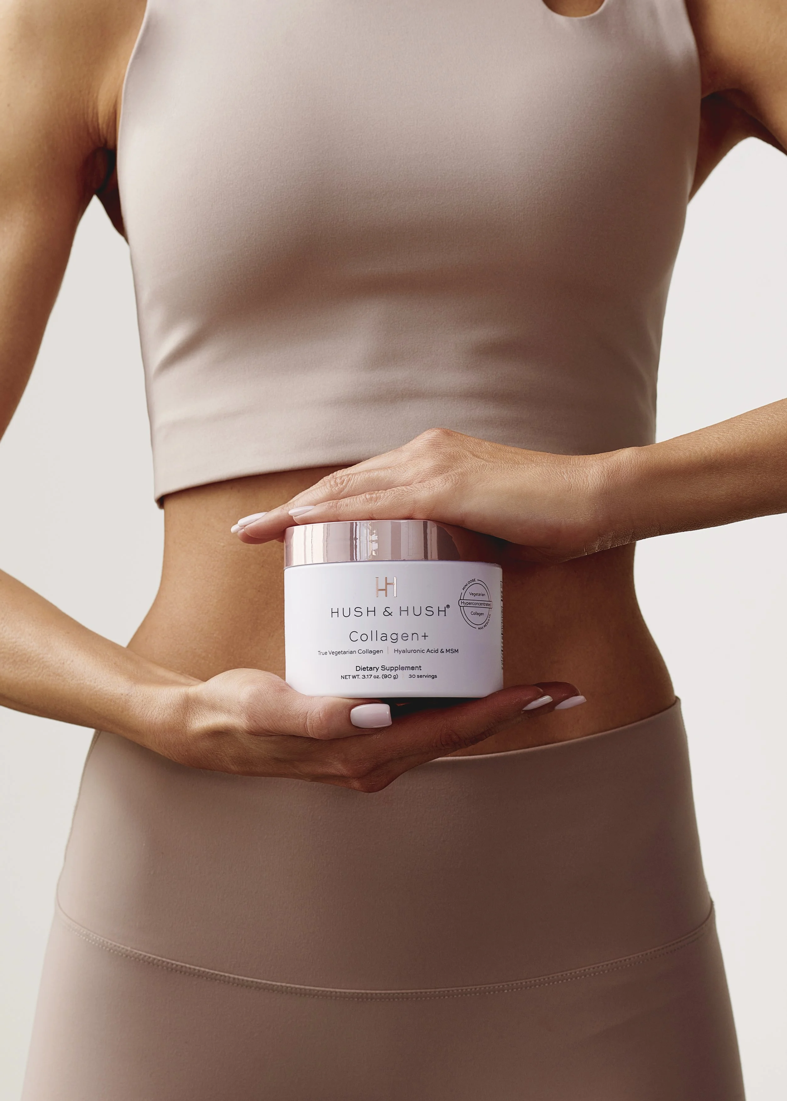 Person holding Hush & Hush Collagen+ dietary supplement in front of their abdomen, wearing beige activewear.