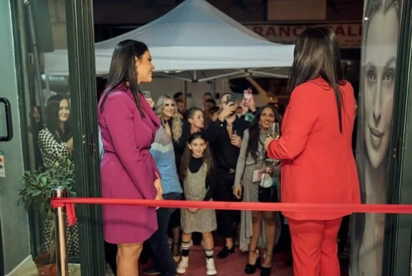 Two women at ribbon-cutting ceremony with crowd in background, one in red suit and the other in purple dress, outside a venue with a white canopy.