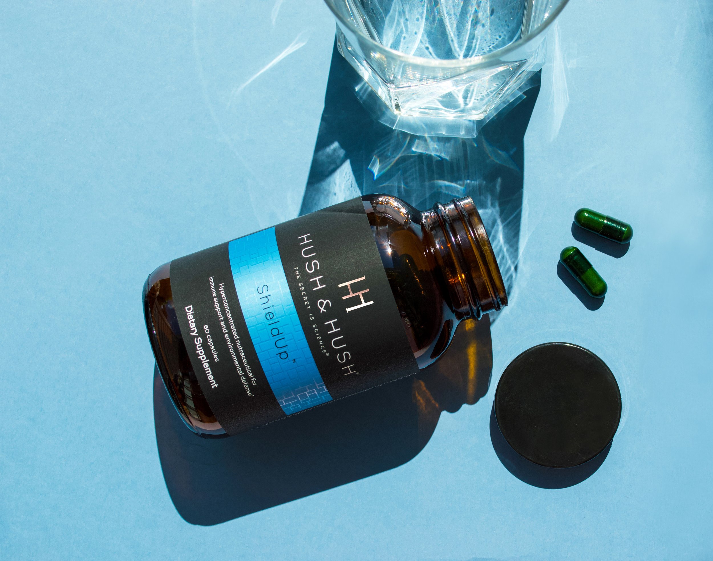 Hush & Hush ShieldUp dietary supplement bottle on blue background with capsules and glass of water.