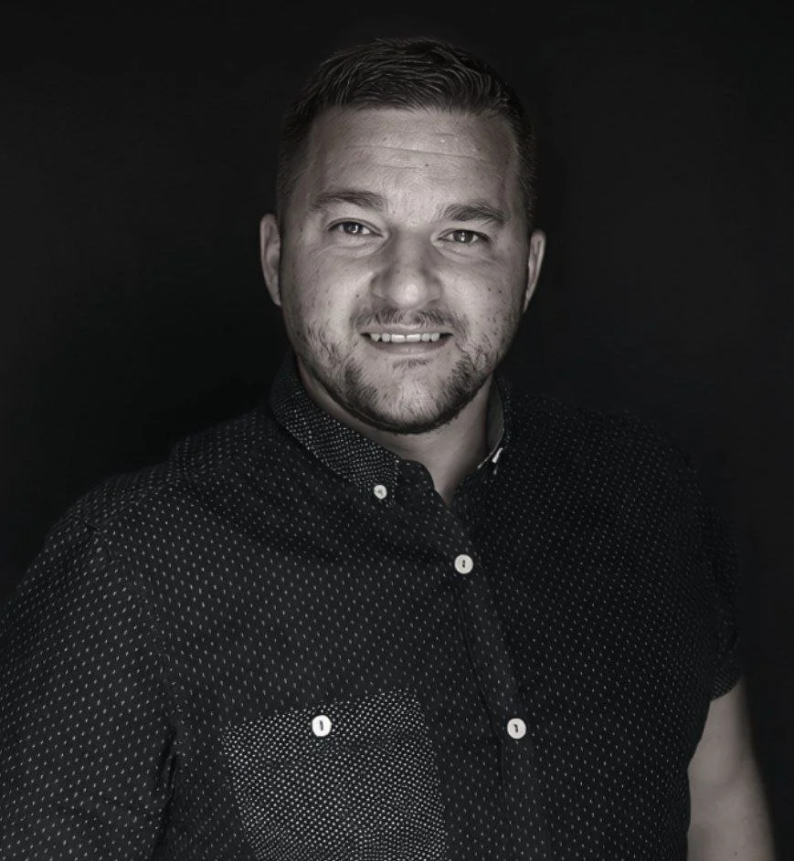 Black and white portrait of a young man with short hair, light beard, wearing a dark patterned button-up shirt, smiling slightly, against a dark background.