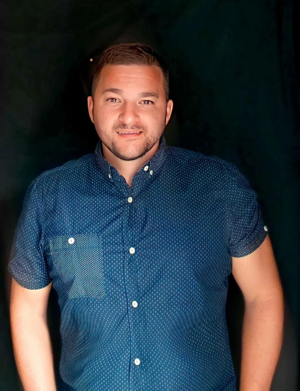 John Balding with short brown hair, a beard, and a light complexion, wearing a navy blue short-sleeved button-up shirt with small white polka dots, standing against a dark background.