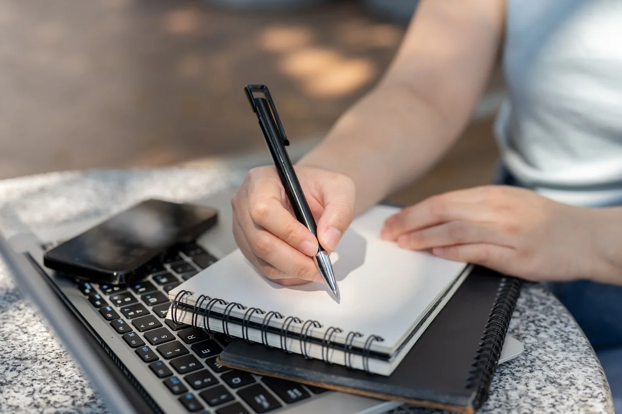 Person writing in a spiral notebook with a pen, placed on a laptop keyboard, with a smartphone nearby on a granite table.