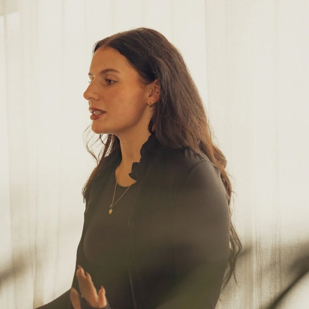 Emily, instructor at Sojourn Pilates in Broughton Astley, Leicestershire, in profile wearing a charcoal zip-up activewear top with delicate gold necklaces, soft daylight through linen curtains behind her.