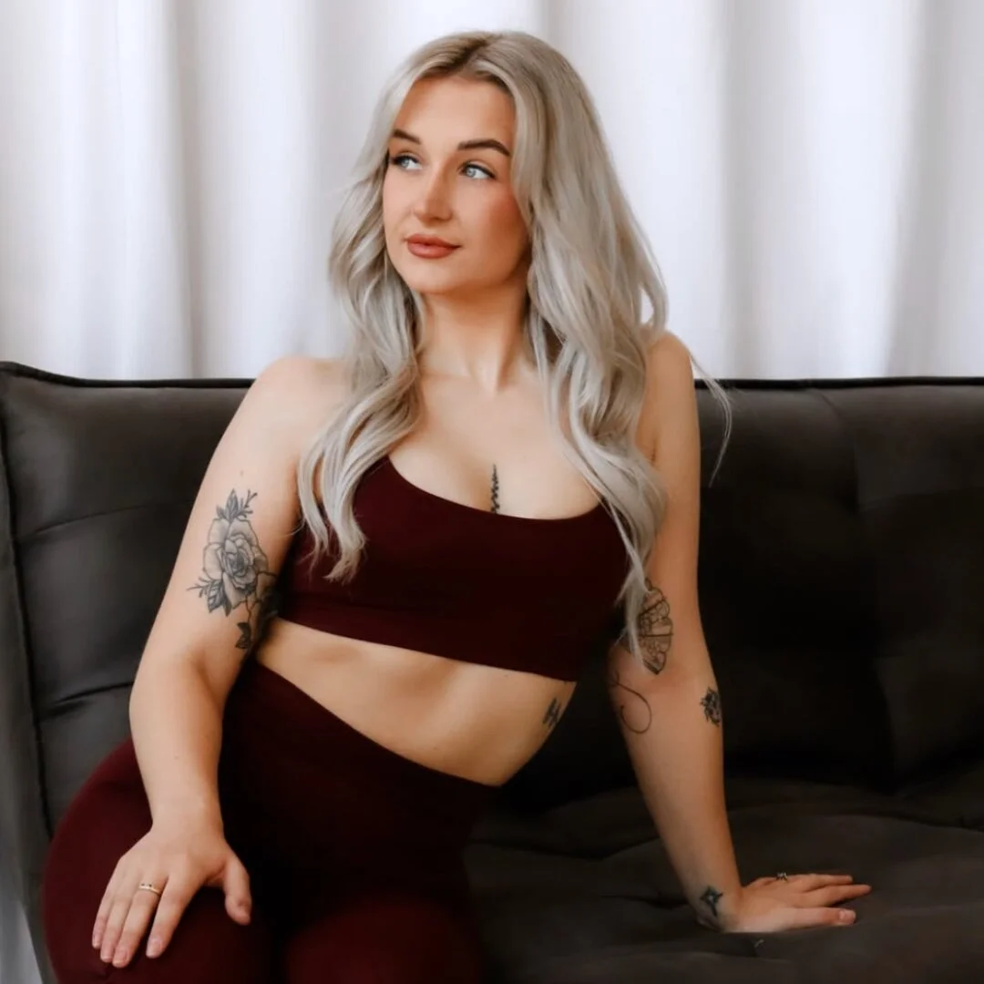 Lorna, instructor at Sojourn Pilates in Broughton Astley, Leicestershire, seated on a dark sofa in a burgundy sports bra and leggings, floral arm tattoos visible against a soft curtain backdrop.