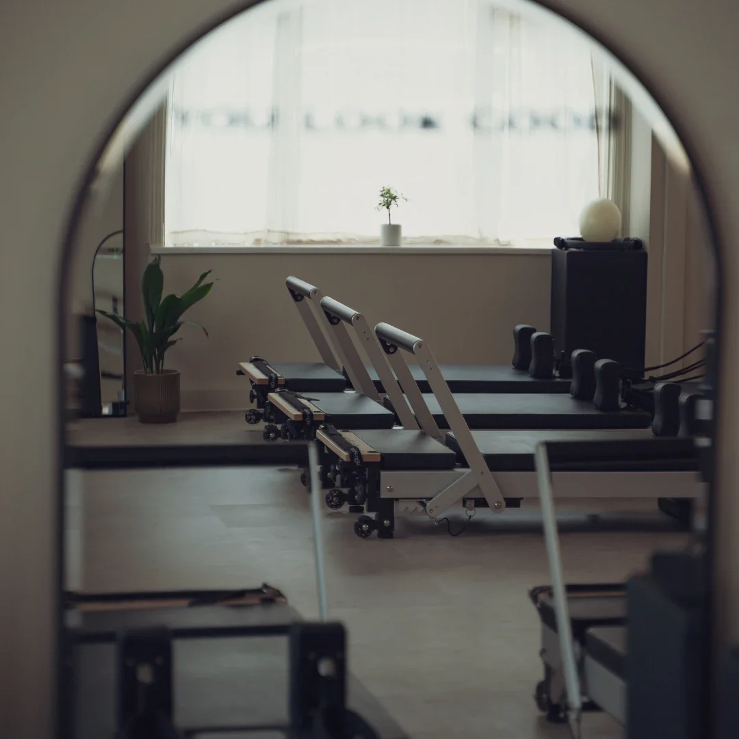 A row of reformer beds at Sojourn Pilates in Broughton Astley, Leicestershire, lit by natural light through linen curtains, with the studio's 'You Look Good' window decal softly in view.
