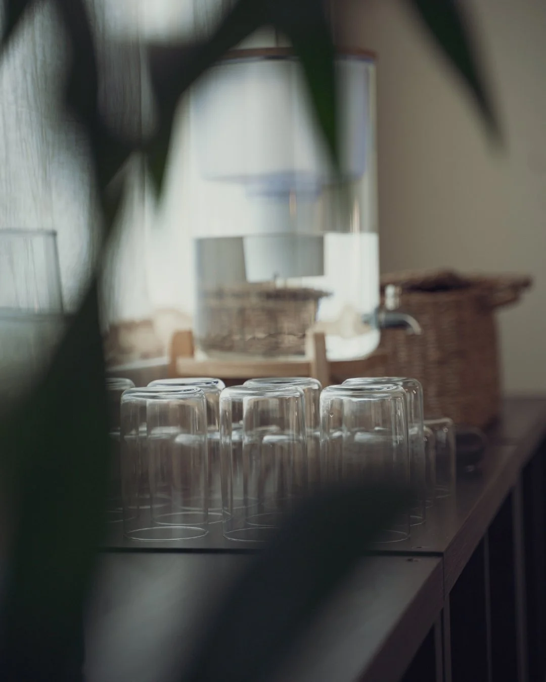 The alkaline water station at Sojourn Pilates in Broughton Astley, Leicestershire, with upside-down glasses lined on a dark wooden counter, a woven basket beside, and soft daylight catching the plants behind.