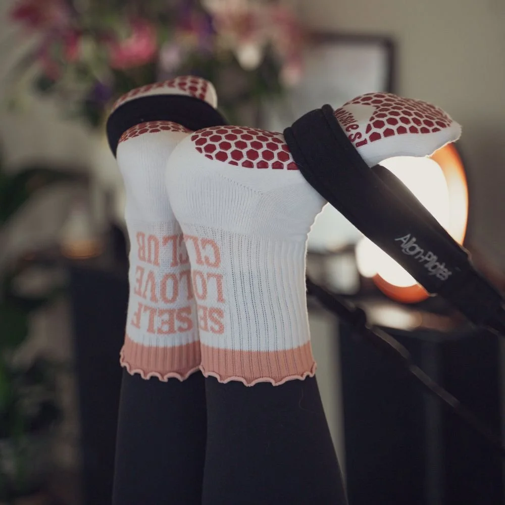Feet in Self Love Club grip socks raised in the reformer straps during a Sojourn Pilates footwork sequence, infrared light warming the studio behind.