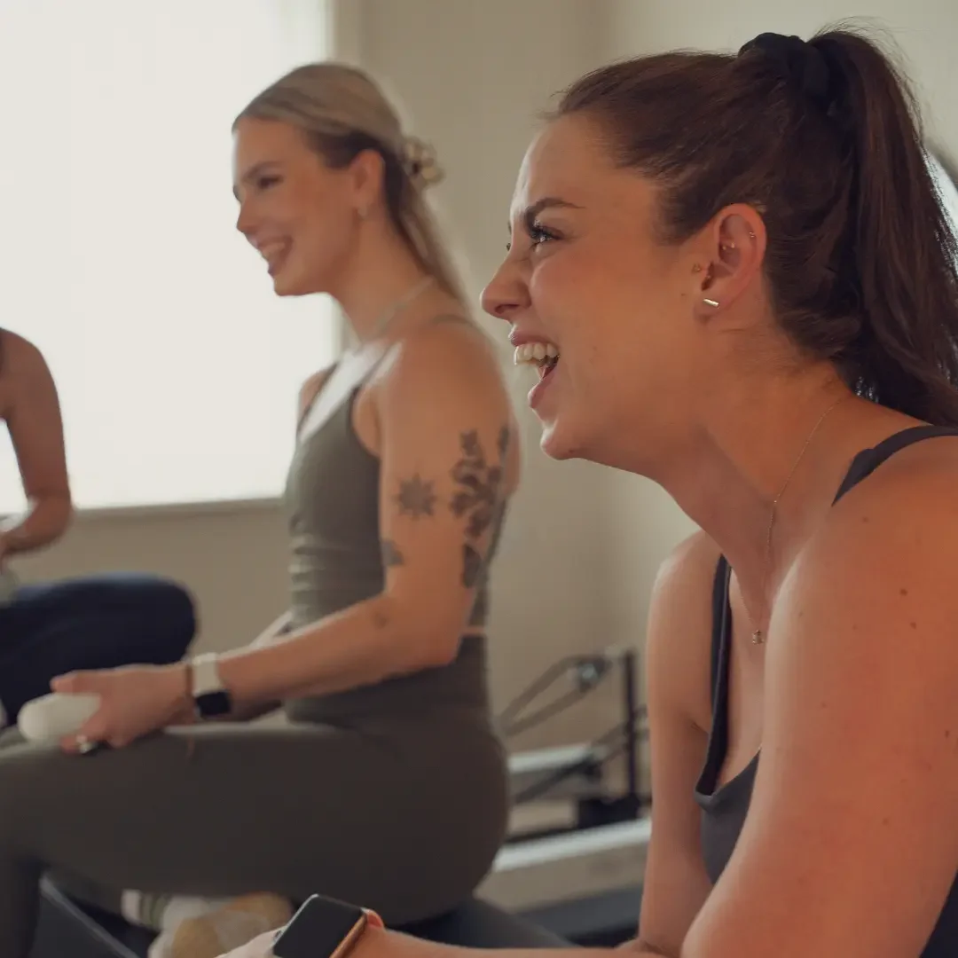 Two friends laughing on the reformers during a private group hire class at Sojourn Pilates, a boutique reformer studio in Broughton Astley, Leicestershire.