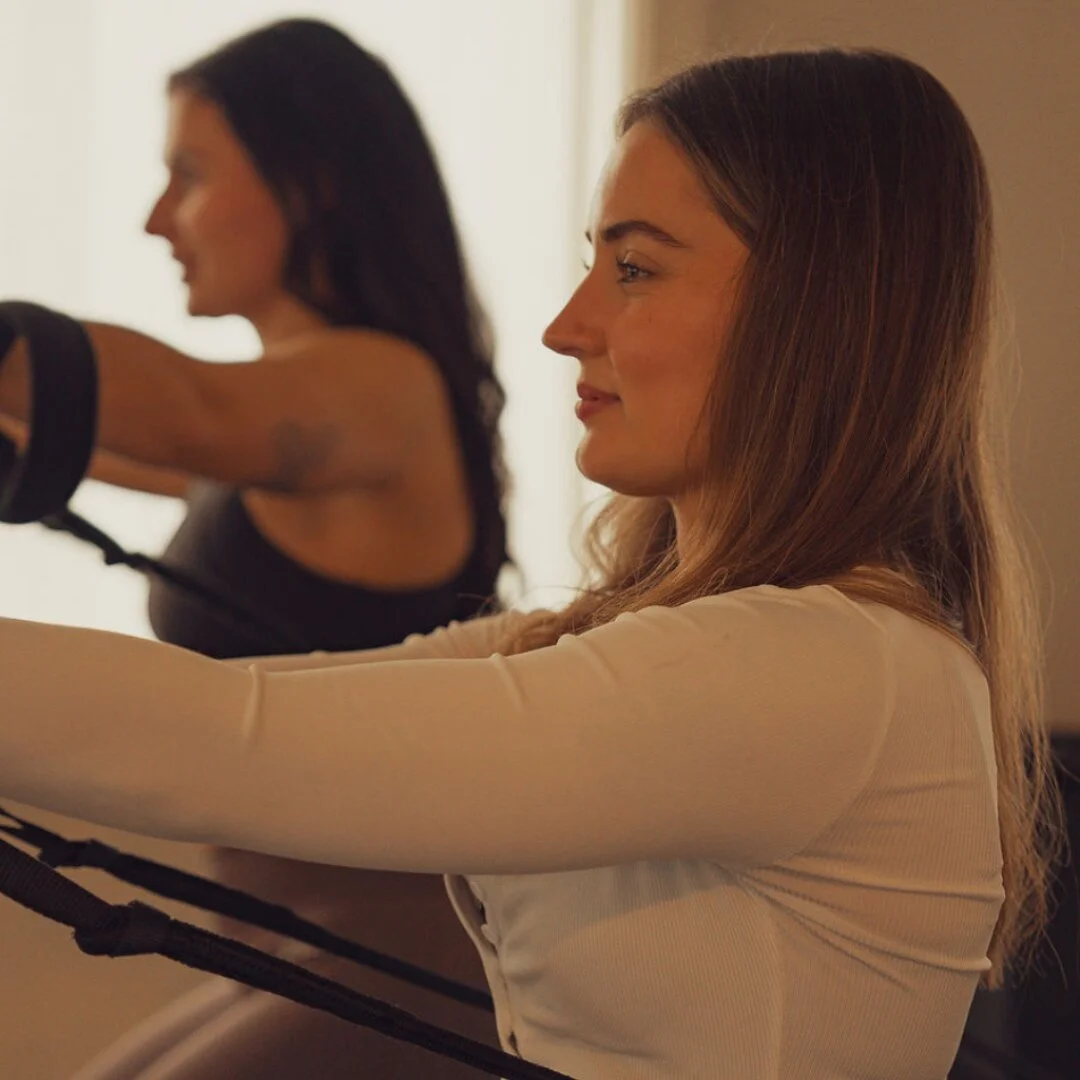 Rachel, Trainee Instructor at Sojourn Pilates in Broughton Astley, Leicestershire, in profile working with reformer straps in a cream long-sleeve top, a fellow client visible beside her in the warmly lit studio.