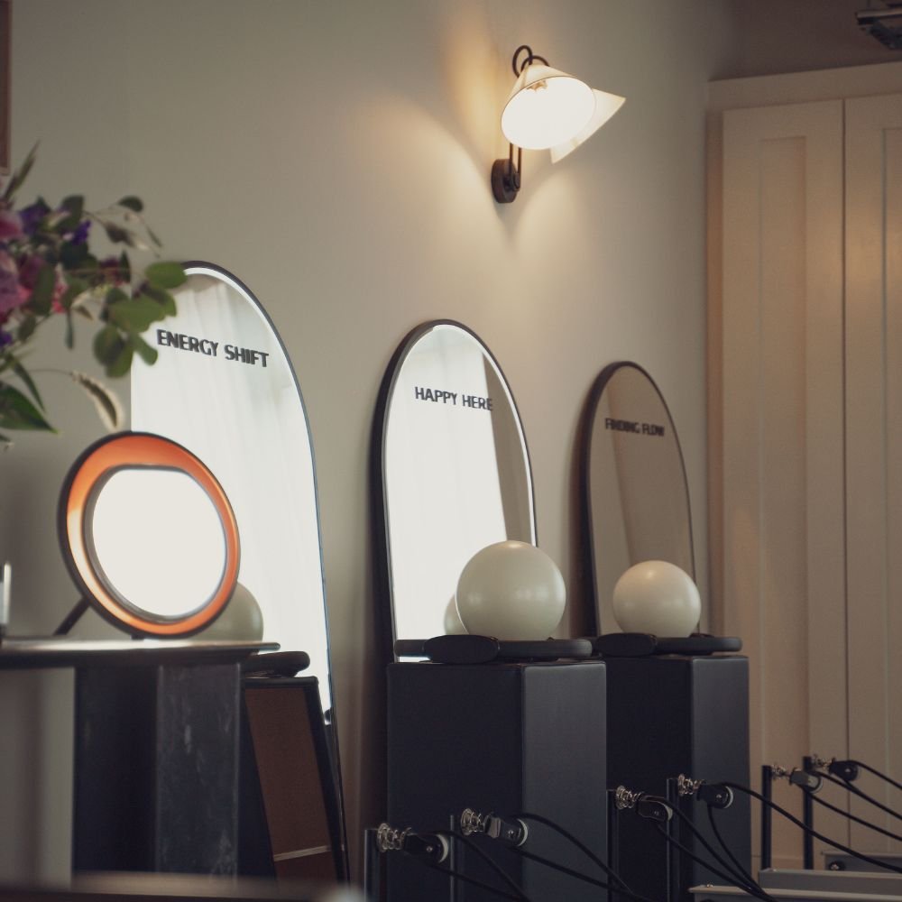 The affirmation mirrors at Sojourn Pilates in Broughton Astley, Leicestershire, printed with 'Energy Shift' and 'Happy Here', framed by a warm wall sconce and seasonal flowers.