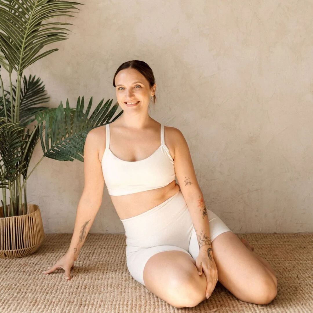 Hayley, instructor at Sojourn Pilates in Broughton Astley, Leicestershire, smiling in a cream Pilates set as she sits gracefully on a woven jute rug beside a potted palm, floral tattoos along her forearms and a dancer's poise in her resting pose.