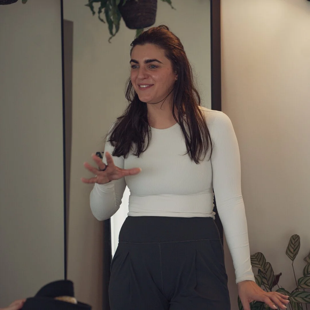 Charlotte, founder of Sojourn Pilates, mid-conversation in a cream long-sleeve top inside the Broughton Astley studio in Leicestershire, with a hanging plant and infrared affirmation mirror behind her.