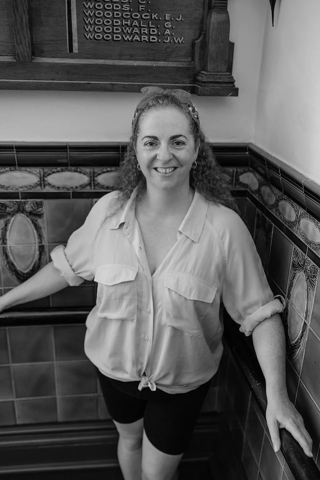 A woman with curly hair wearing a tied-up light-colored shirt and dark shorts, smiling, standing in a tiled stairwell with decorative tiles on the wall.