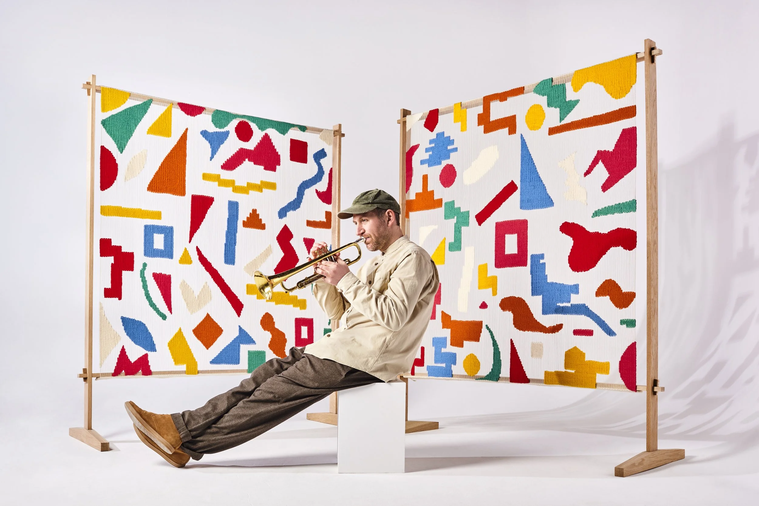 A man sitting on a white block playing a trumpet in front of two colorful geometric art panels on a white background.