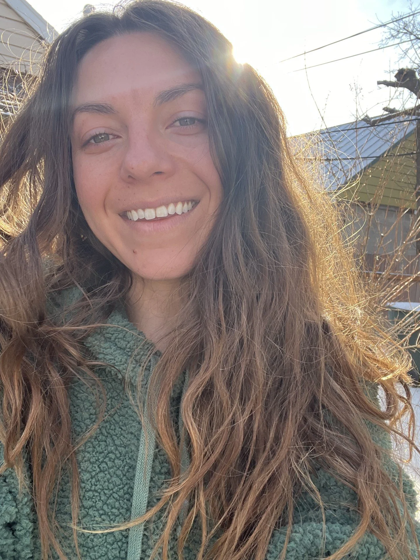 Close-up of a smiling woman with long, wavy hair outdoors on a sunny day, with a house and tree in the background.