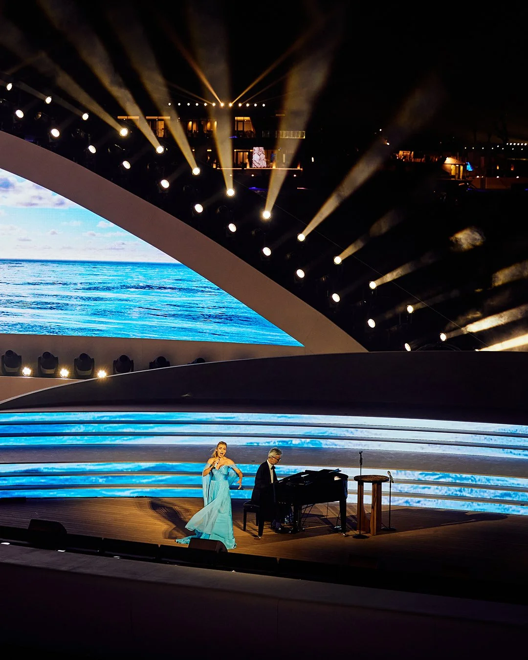 A singer performing on stage with a pianist, during a concert at night with ocean-themed background visuals and stage lighting.