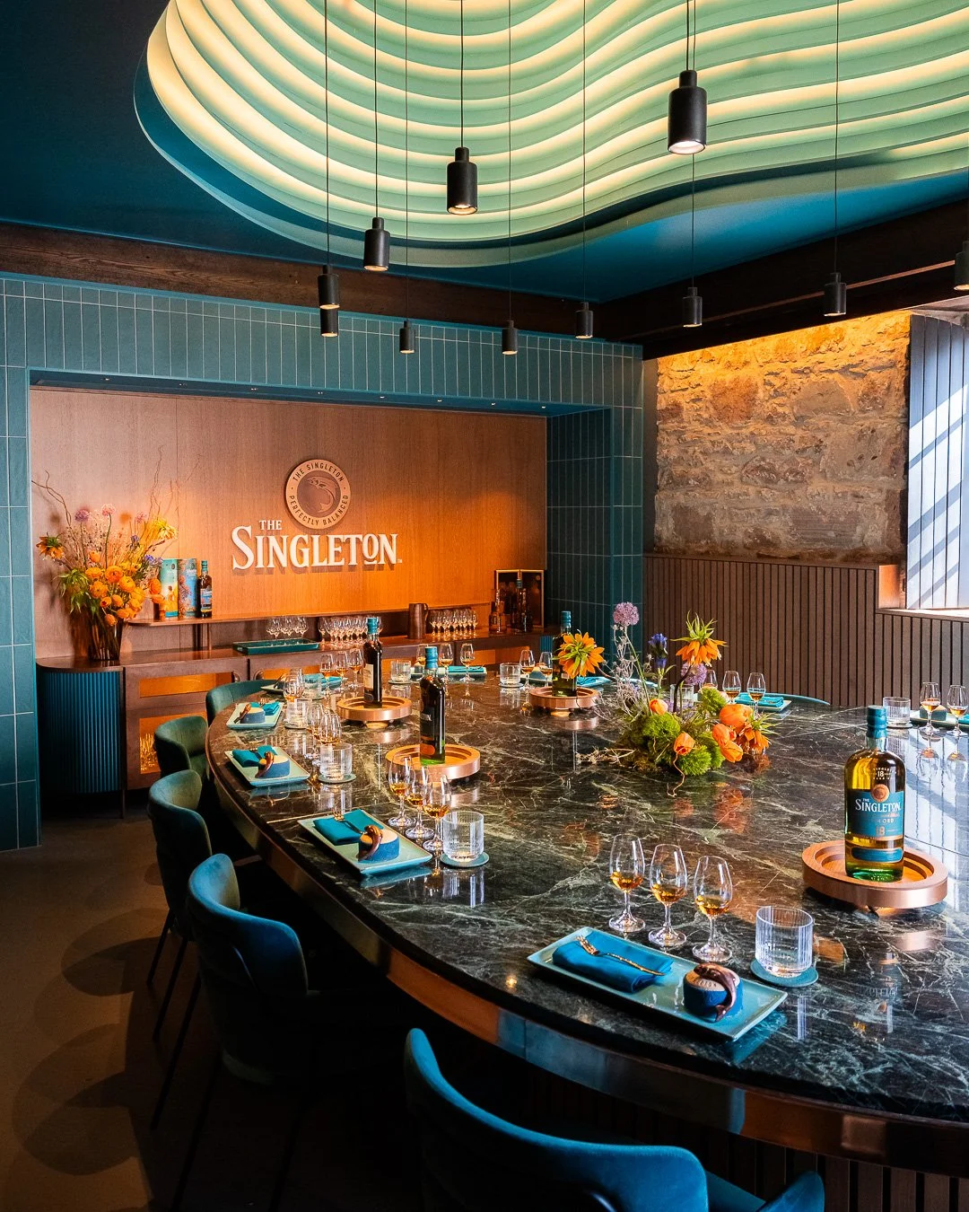 A stylish dining room with a large, oval black marble table set with glasses, plates, and napkins, with colorful floral centerpieces. The room has a green ceiling with modern lighting, teal tiled walls, a textured stone wall, and a wooden backdrop di