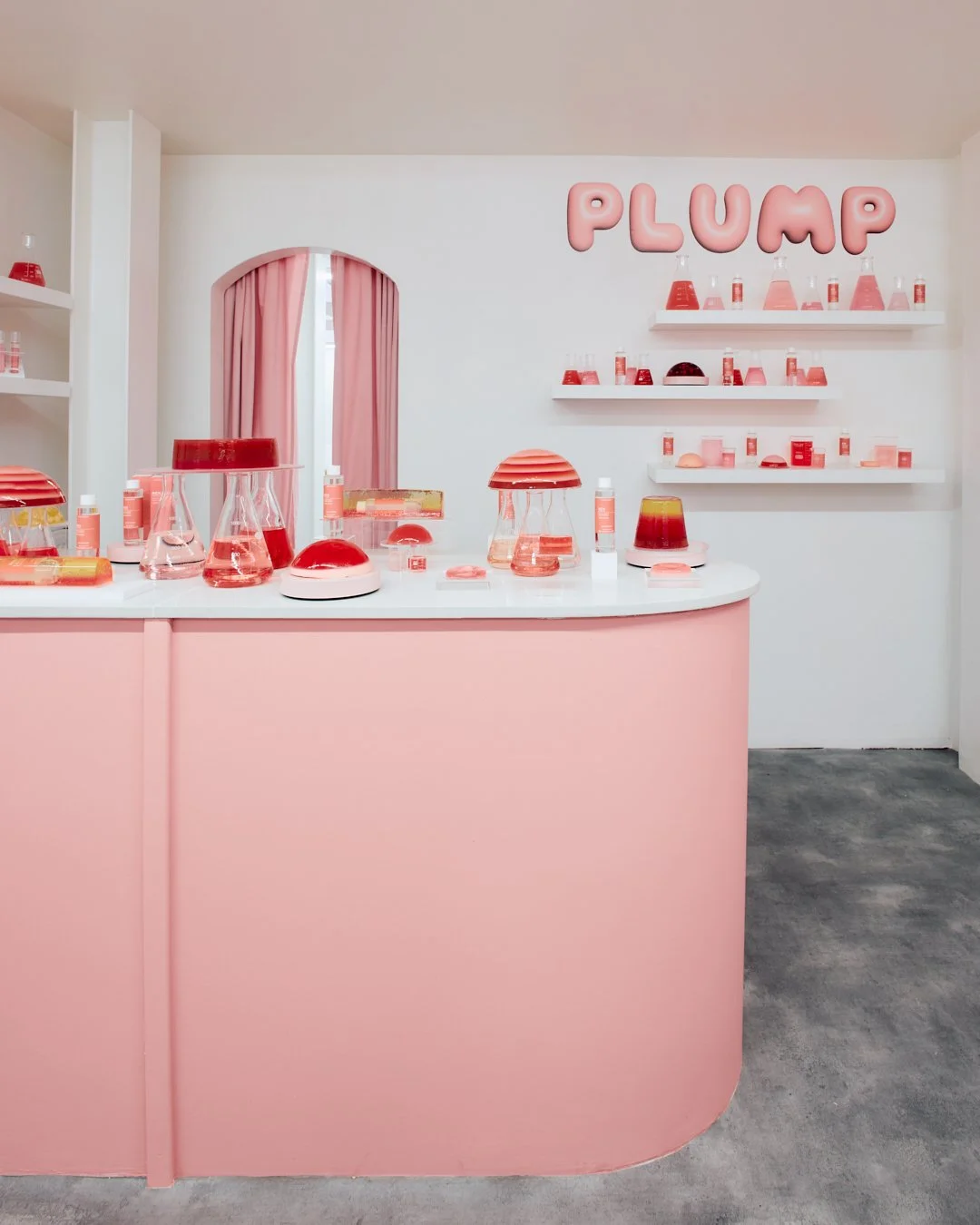 A pastel pink and white display area with various pink and red laboratory glassware and cosmetic bottles, a small mirror with pink curtains, and pink balloon-style letters spelling 'PLUMP' on the wall.