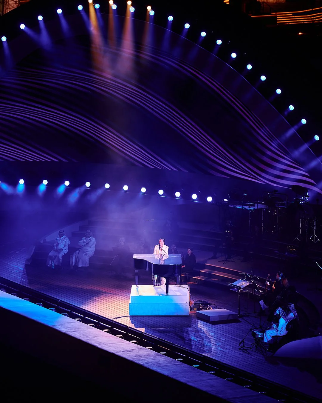 Performer singing at a grand piano on a stage with blue and purple lighting, with backup musicians and dancers in the background, and an elaborate curved light setup overhead.