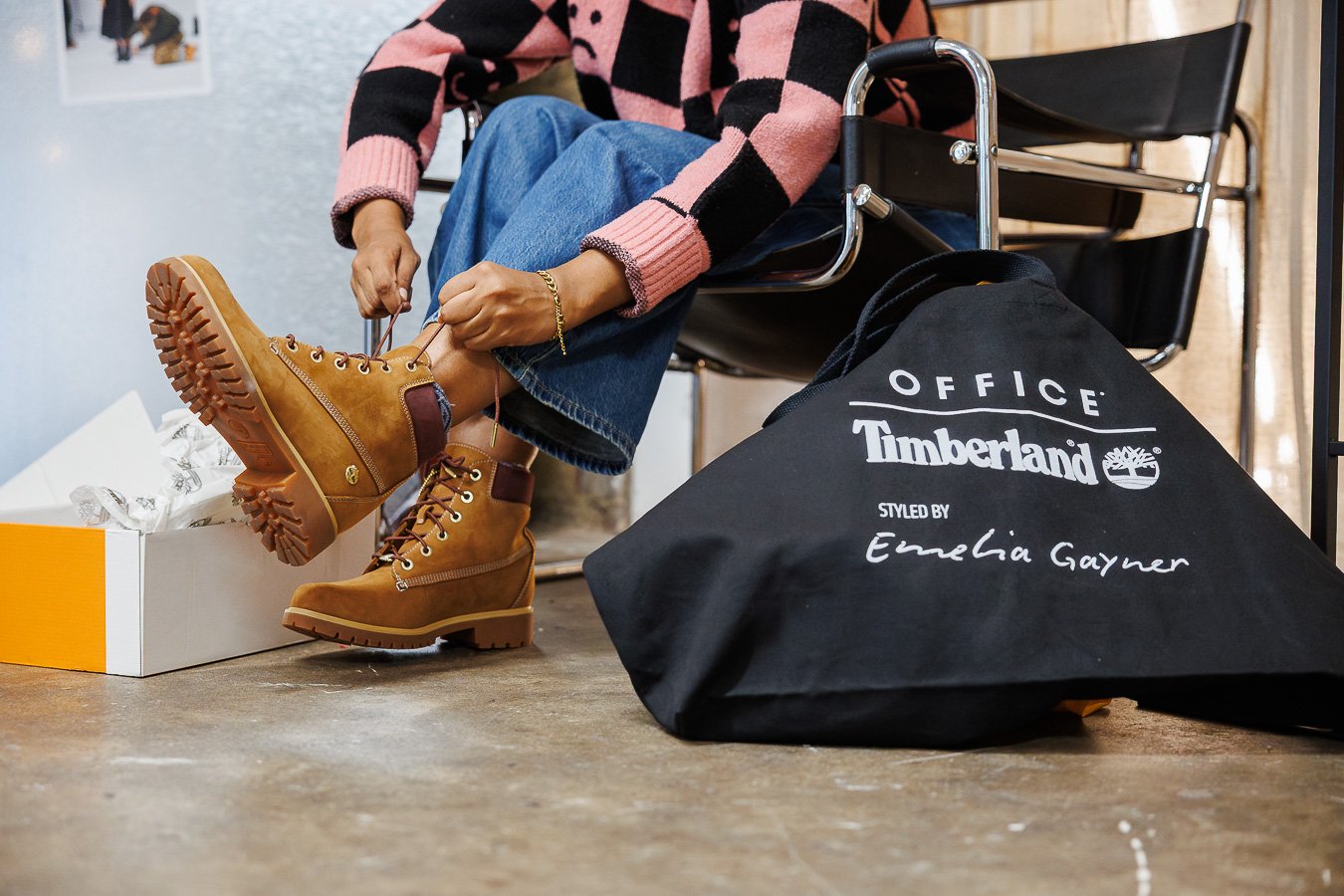 Person sitting on a black chair, tying brown Timberland boots, with a black Timberland shopping bag on the floor nearby.
