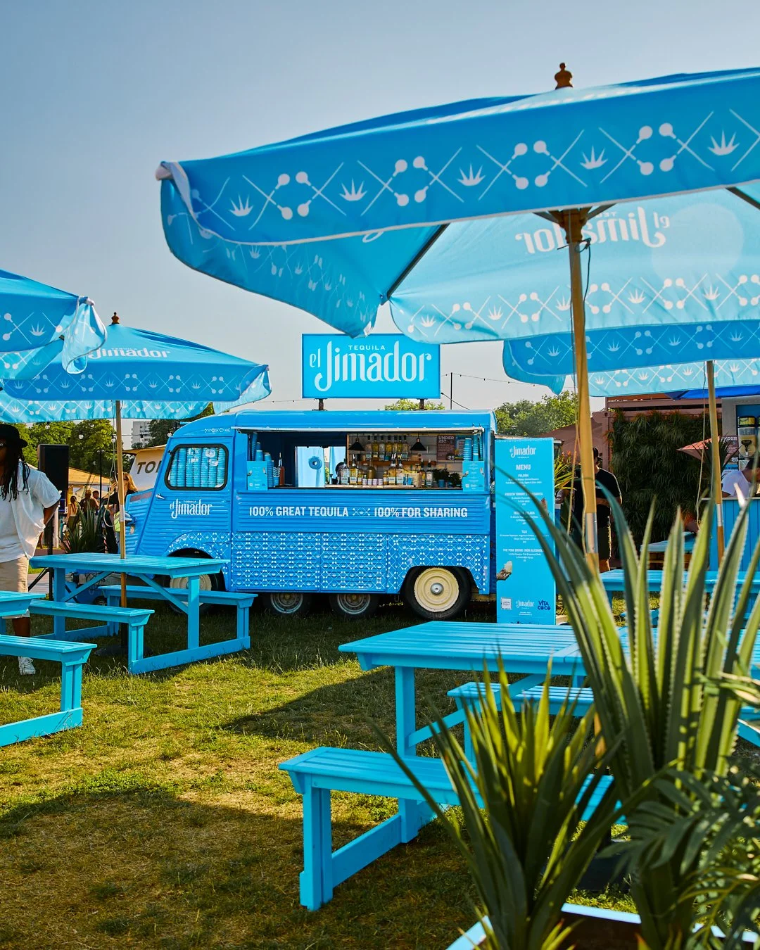 Bright blue mobile tequila bar with a sign that reads "El Jimador" at an outdoor event, surrounded by blue tables, large umbrellas, and greenery.