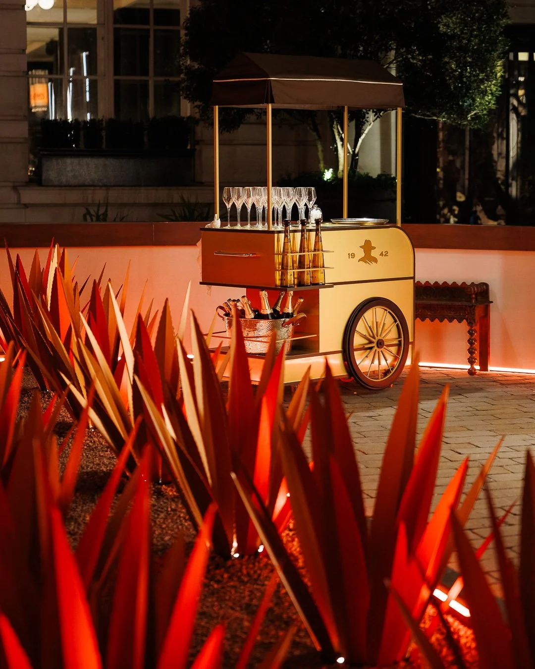 A vintage-style bar cart with a small umbrella, glasses, bottles, and a decorative hat image, set outdoors at night with warm lighting, surrounded by plants and a stone or brick paved surface.