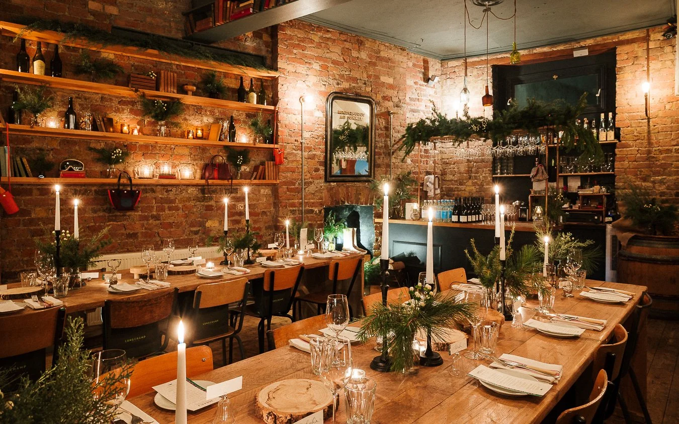A cozy, rustic restaurant decorated for the holidays with wooden tables, candlelit candles, and greenery. Exposed brick walls feature shelves with bottles, books, and decorative items. The setting includes neatly arranged glassware and plates for din