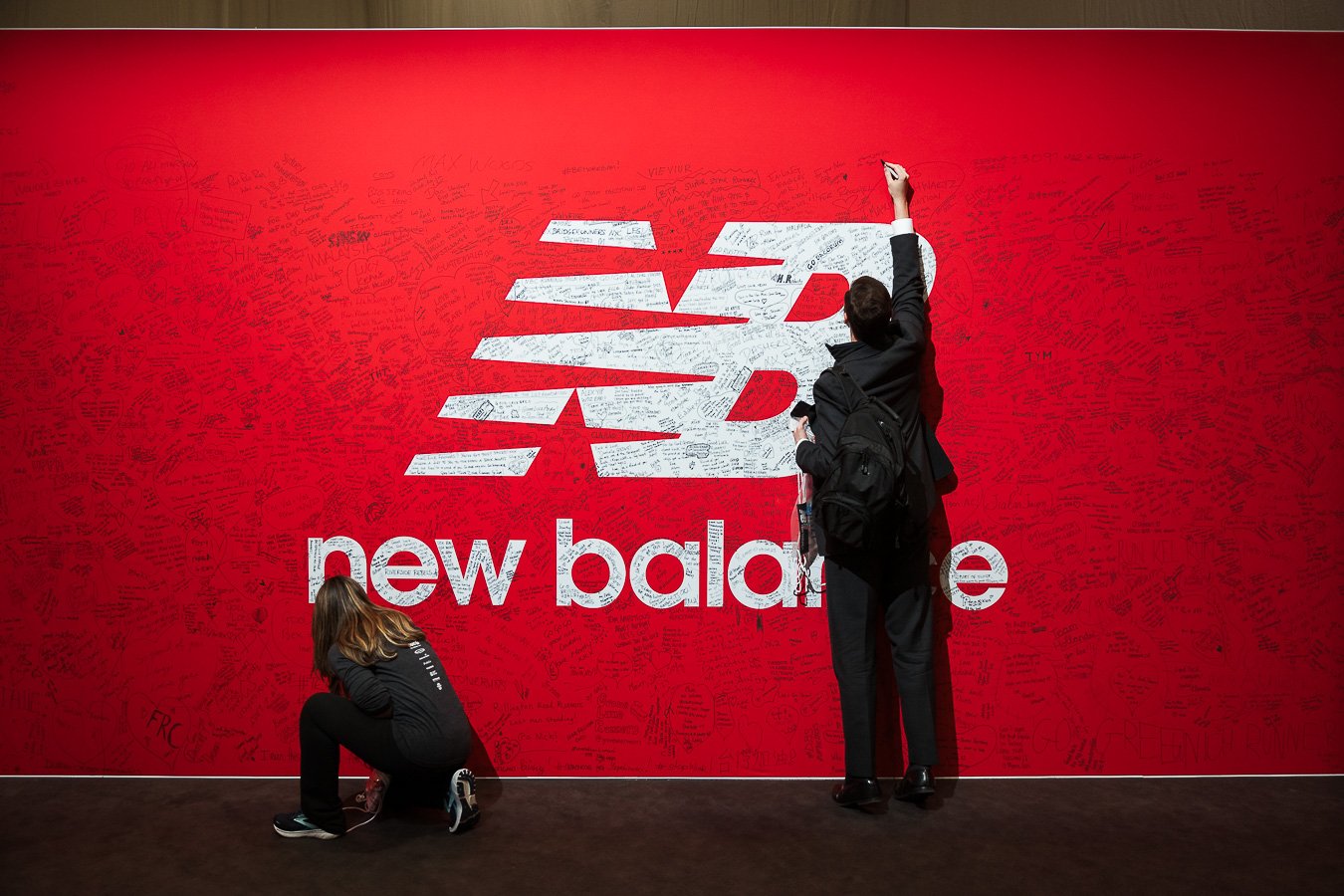 Two people signing a large red wall with the New Balance logo, which is filled with handwritten signatures and messages.
