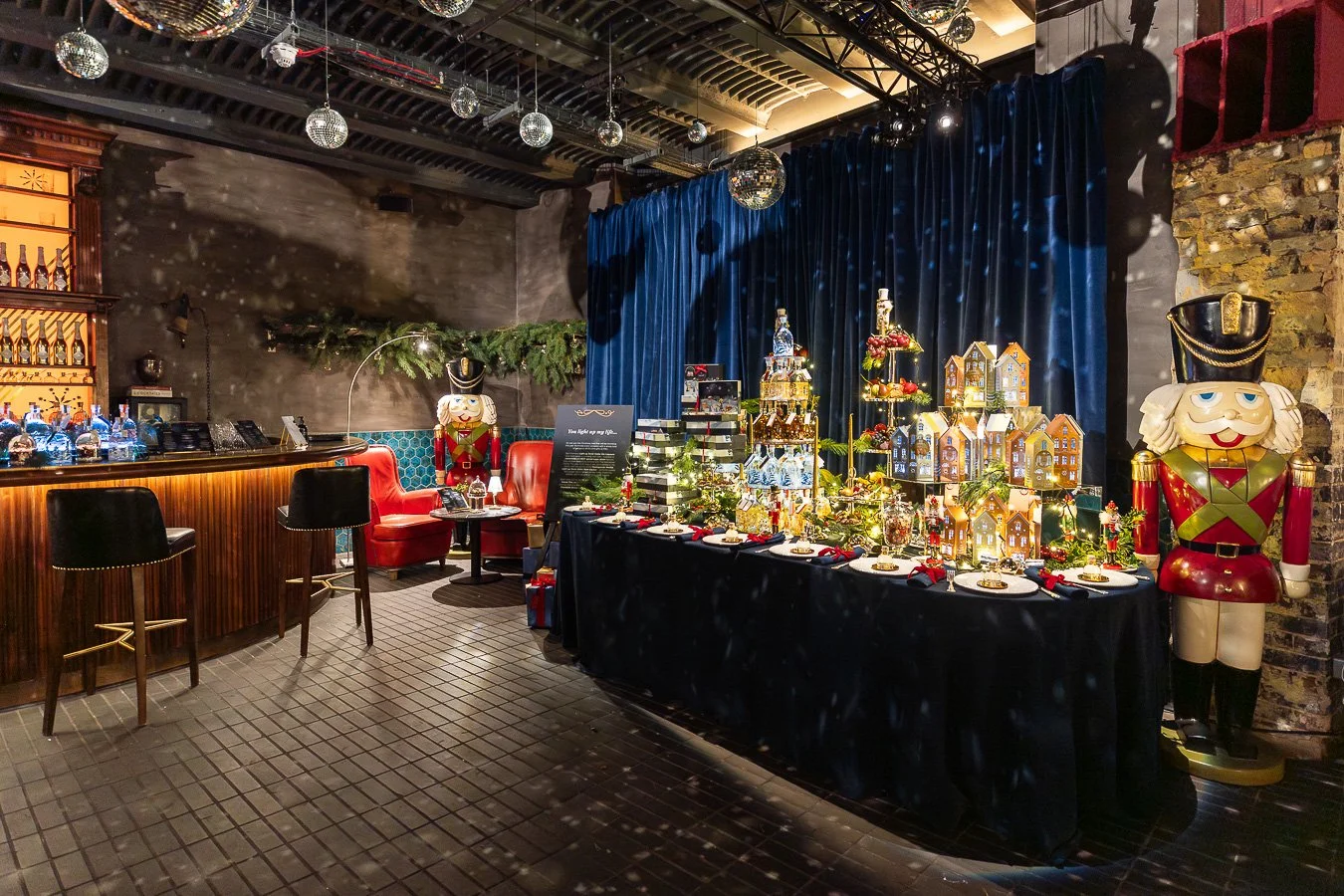 A festive holiday interior with a decorated table of gingerbread houses, nutcracker statues, and holiday ornaments. There is a bar on the left with bottles and a seating area with red chairs and black barstools. The background has a dark blue curtain