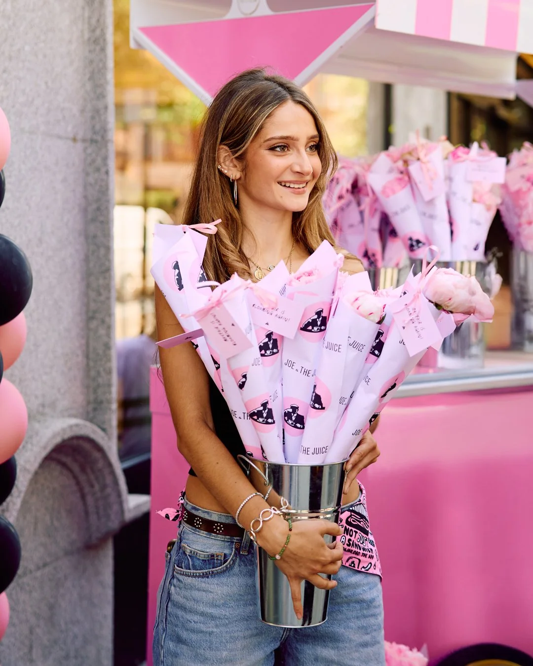 A young woman holding a large bouquet of pink-wrapped flowers, standing outside a shop decorated with pink and black balloons and pink decorations.