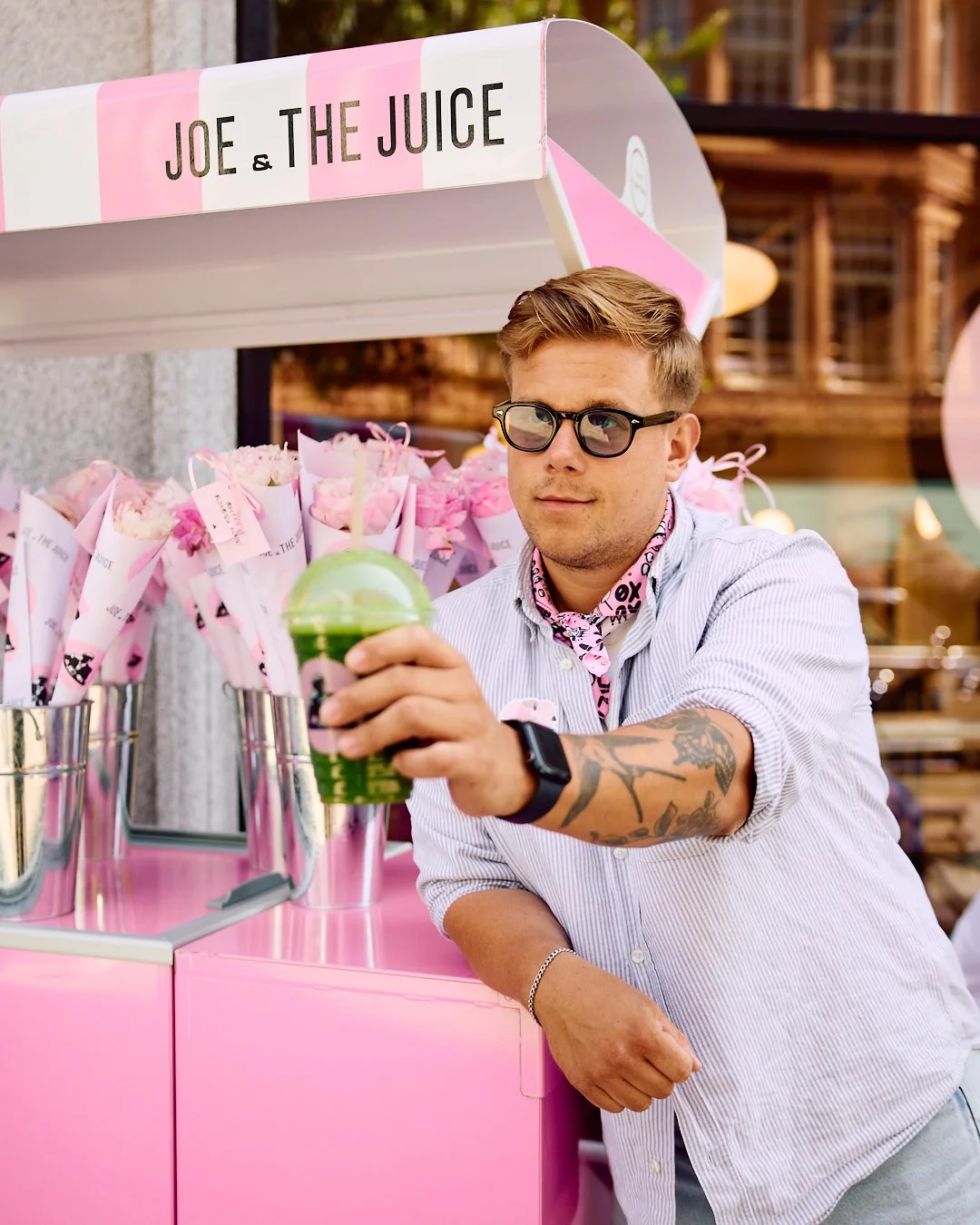 A man with glasses and a tattoo on his arm wearing a pink bandana around his neck is holding a green drink at a pink juice stand named 'Joe & The Juice'.