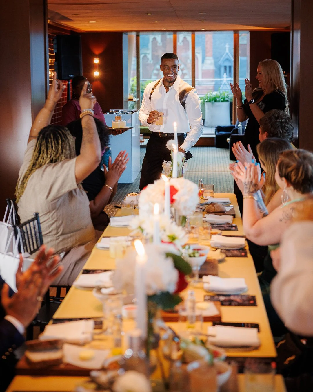 People at a celebratory gathering, clapping and smiling as a man with a backpack enters a room decorated with candles and flowers.