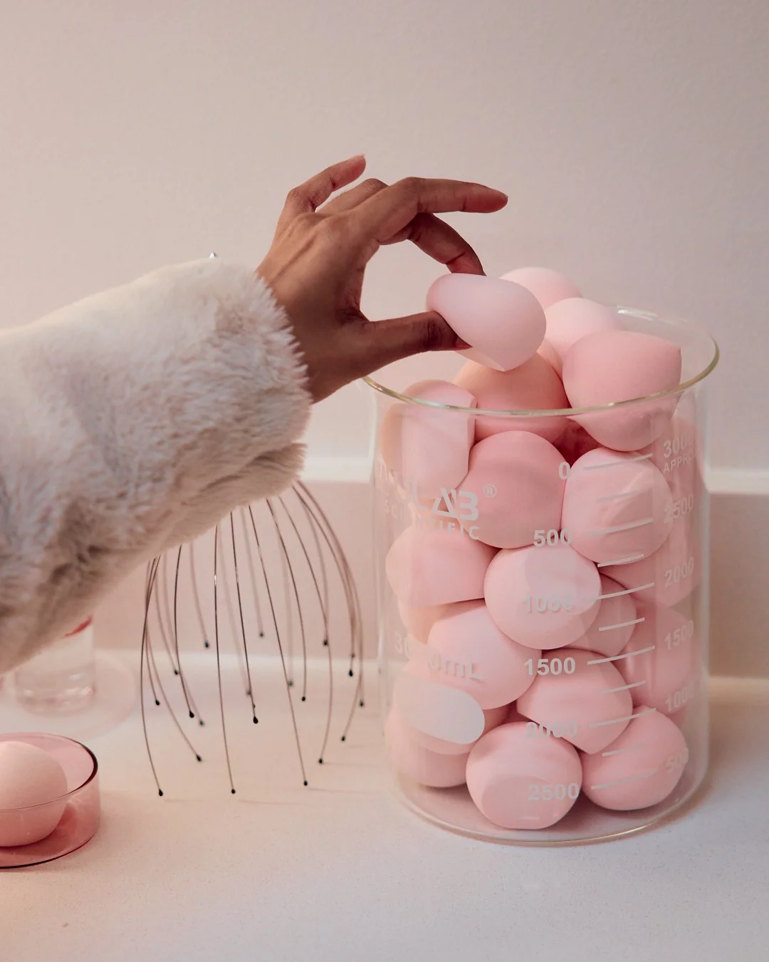 Hand reaching into a large glass measuring cup filled with pink eggs, with a whisk and a small bowl nearby on a white surface.