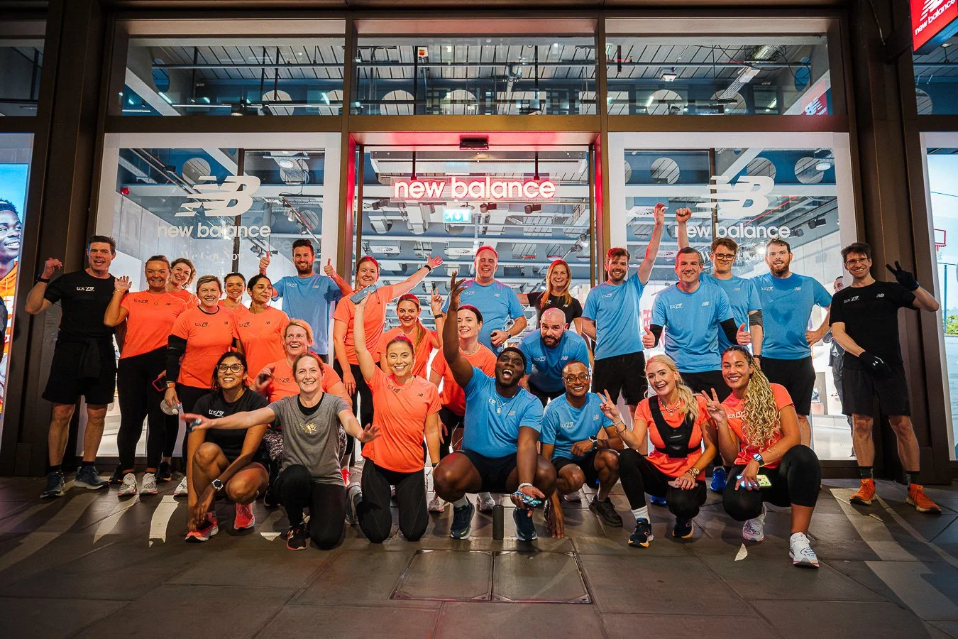 Group of smiling runners in front of a New Balance store, some wearing orange shirts and others in blue, celebrating after a race or event.