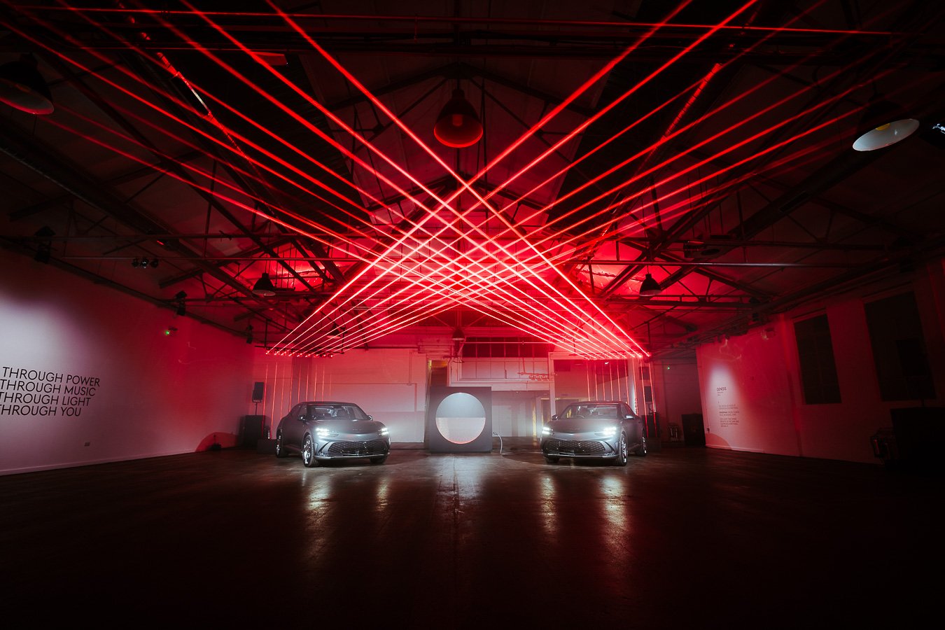 An indoor event space with red laser light beams crossing the ceiling and walls, two cars parked at the front, and digital screens in the background.