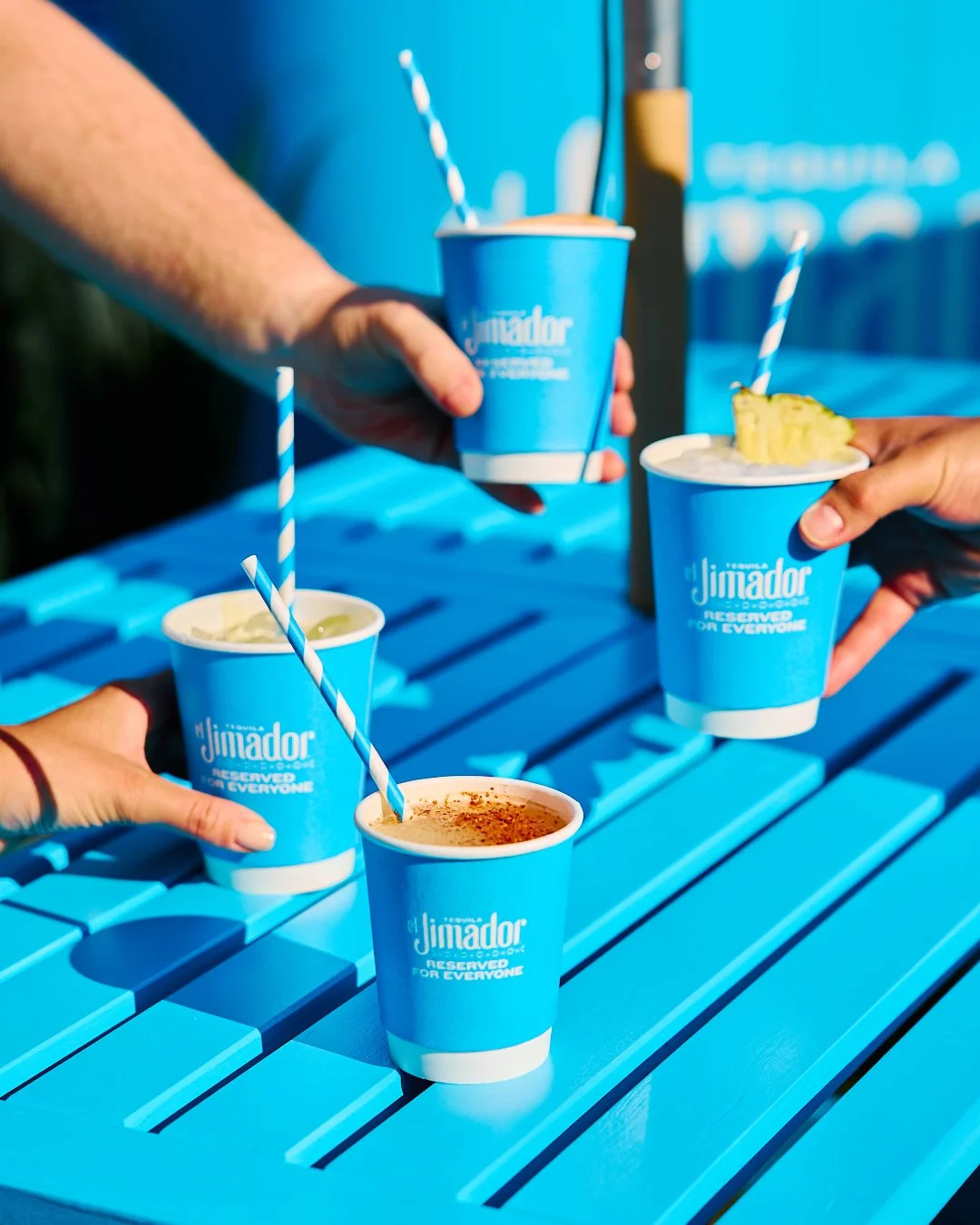 Four people holding blue cups with straws, ready to enjoy frozen beverages or smoothies. The cups have the word 'Jimador' printed on them, and they are placed on a matching blue table.