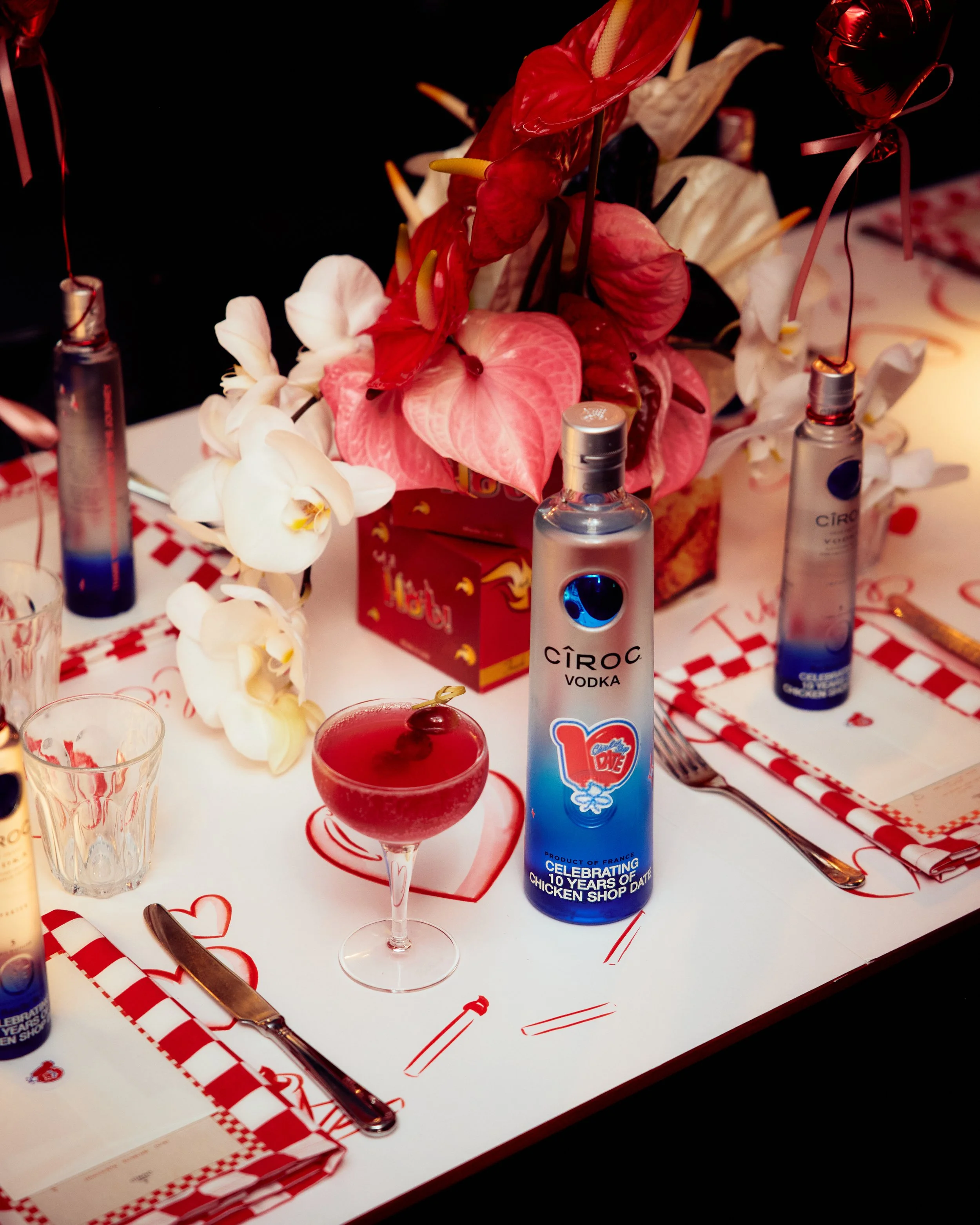 Table setting with a bouquet of pink and white flowers, a bottle of Ciroc vodka with a sticker celebrating 10 years of chicken shop date, a cocktail glass with a red drink and cherry, and heart-themed decorations for a romantic celebration.