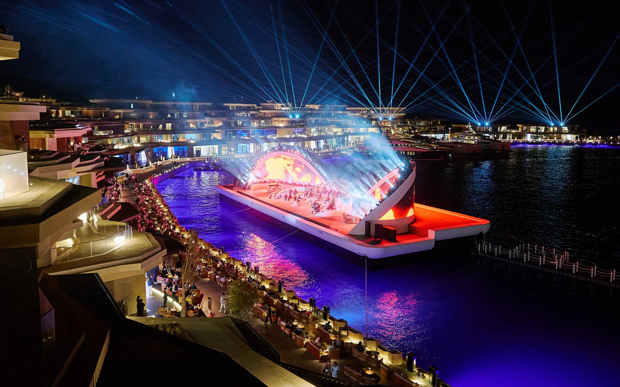 Nighttime view of an outdoor concert stage with colorful lighting, situated on a water body, with audience seating and a modern residential or hotel complex with terraces and blue laser lights in the background.