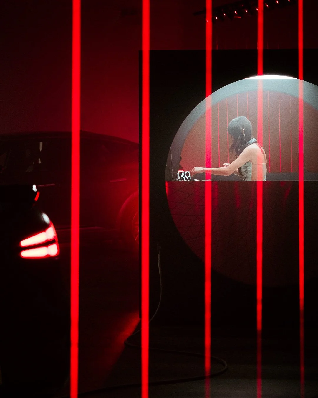 A young woman DJ performing in a dimly lit venue with red lighting, seen through vertical red bars, with a car's taillights visible on the left.